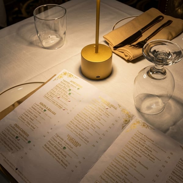 A restaurant table with a menu, golden lamp, empty glass, and silverware on a white tablecloth.